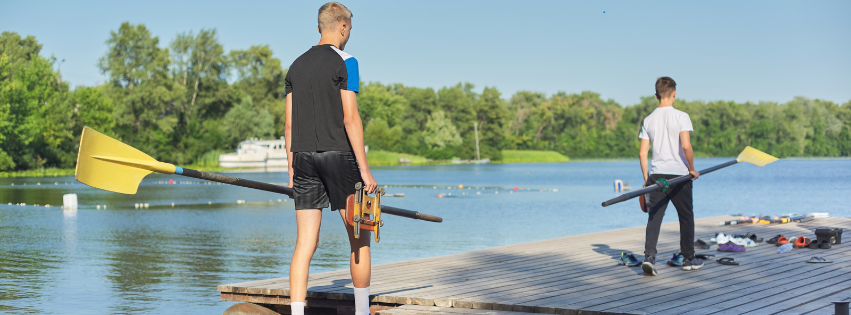 two men hold paddles as they walk toward some paddling gear on a dock