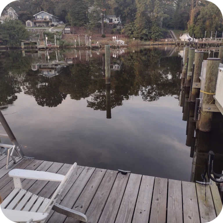 the edge of a dock with a chair in the corner of the photo and trees reflecting on the water at dusk
